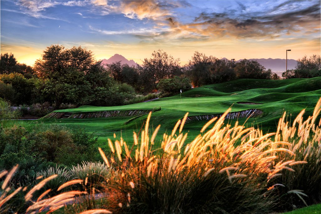 A scenic view of a golf course at sunset with lush green grass, trees, and mountains in the background.