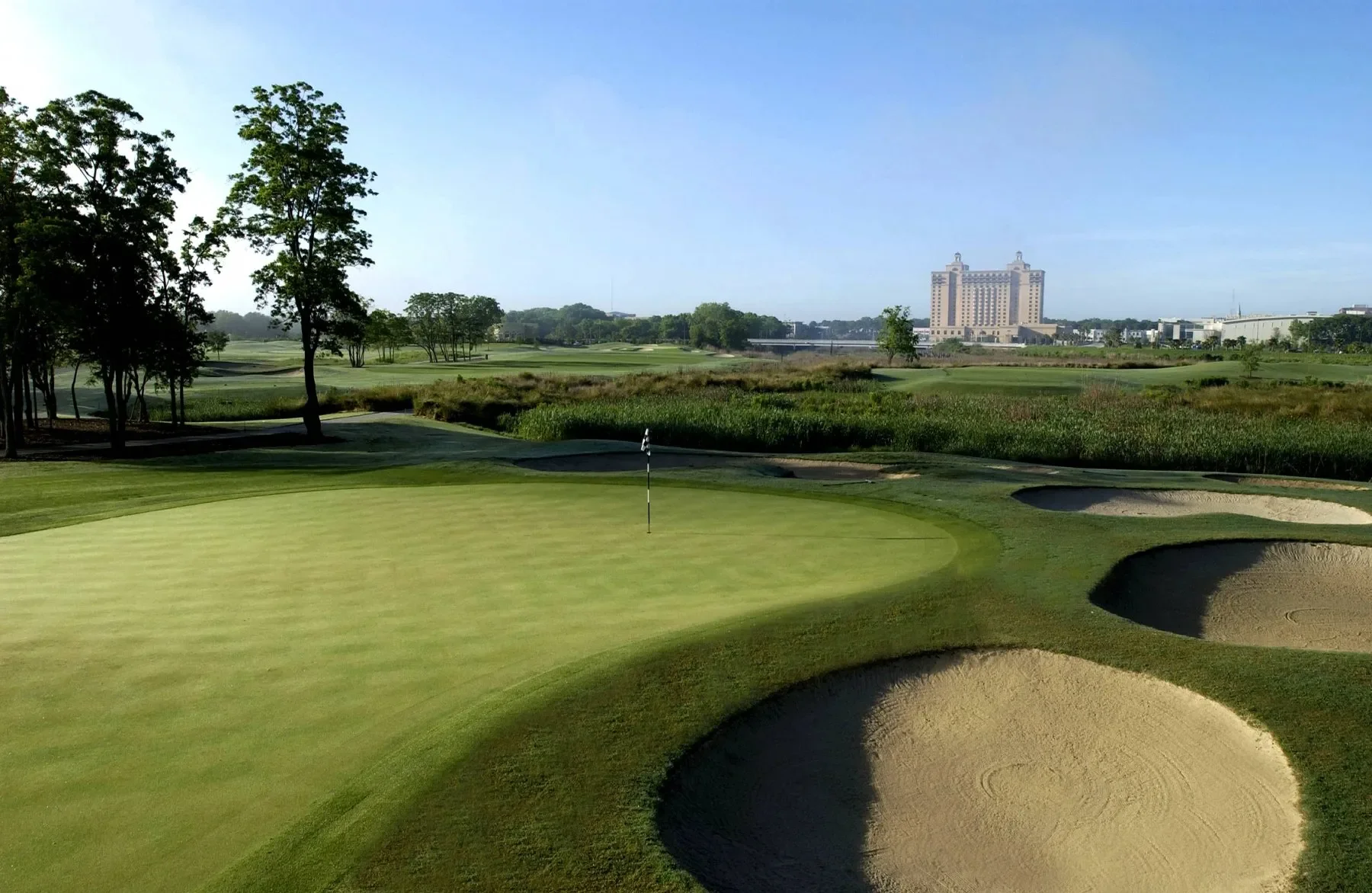 A golf course with a putting green, bunkers, and trees, with a cityscape and a tall building in the background.