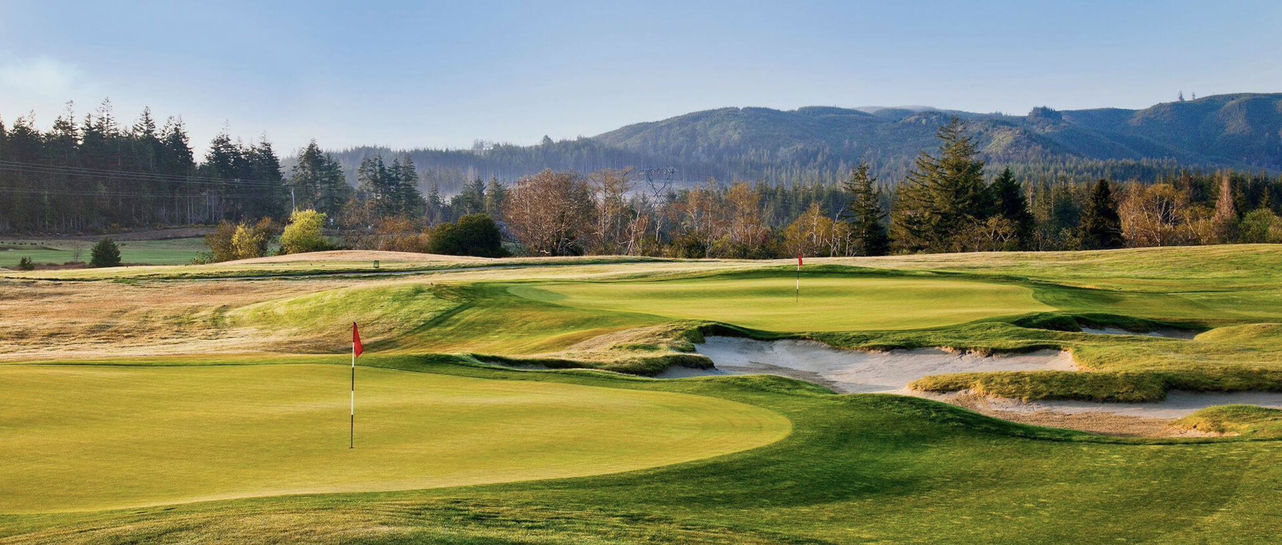 Scenic golf course with green fairways, sand bunkers, and flags, surrounded by trees and mountains in the background.