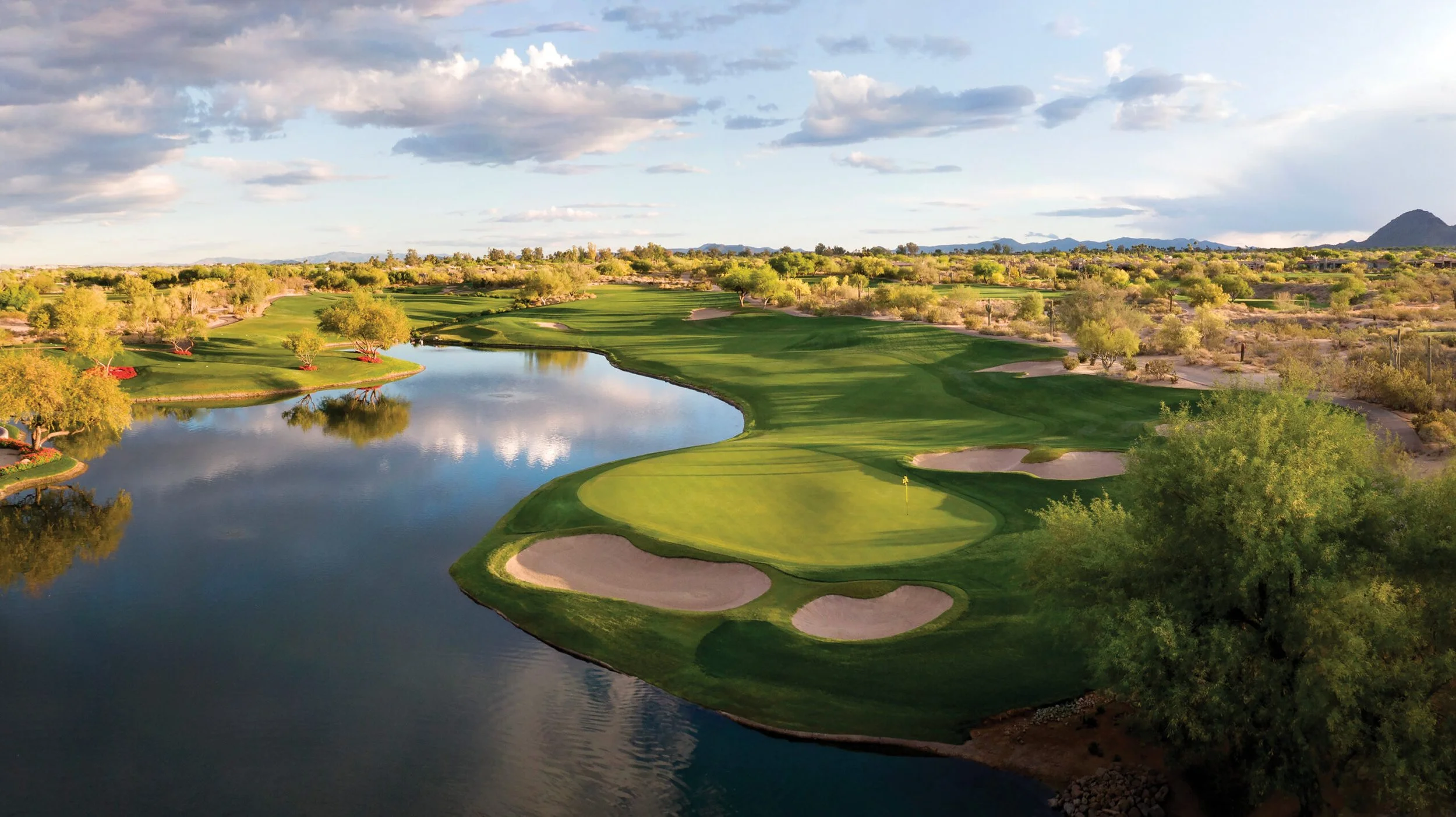 Aerial view of a golf course with green fairways, sand bunkers, a water hazard, and trees under a partly cloudy sky.