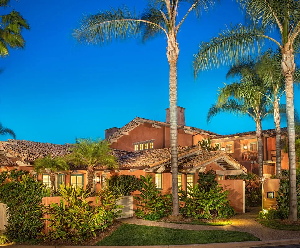 A tropical house with a red exterior, surrounded by tall palm trees and lush green plants, under a clear blue sky at dusk.