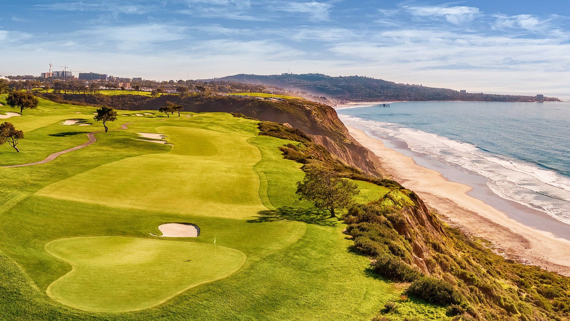 A scenic view of a golf course on a cliff overlooking the ocean with sand dunes, trees, and a city skyline in the background.