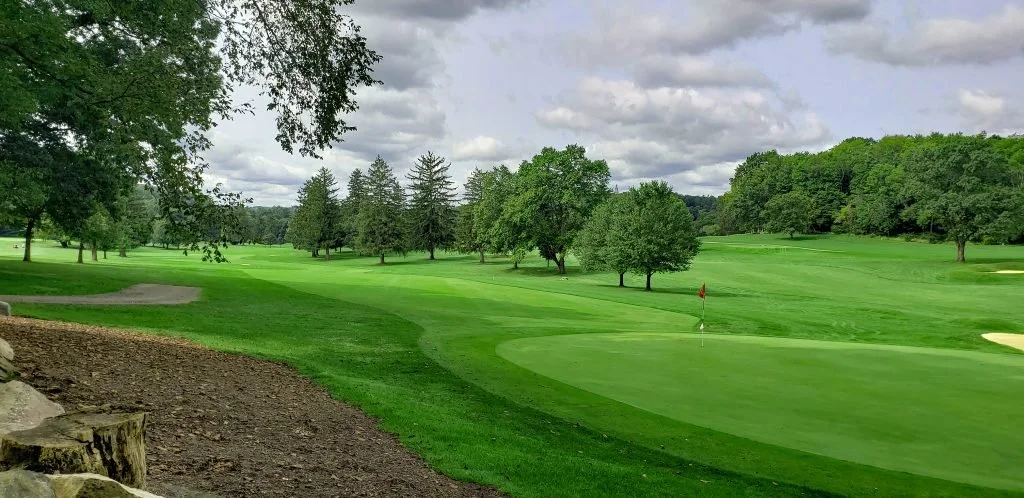 View of a lush golf course with green fairways, a flagstick on the putting green, sand traps, and trees under a partly cloudy sky.