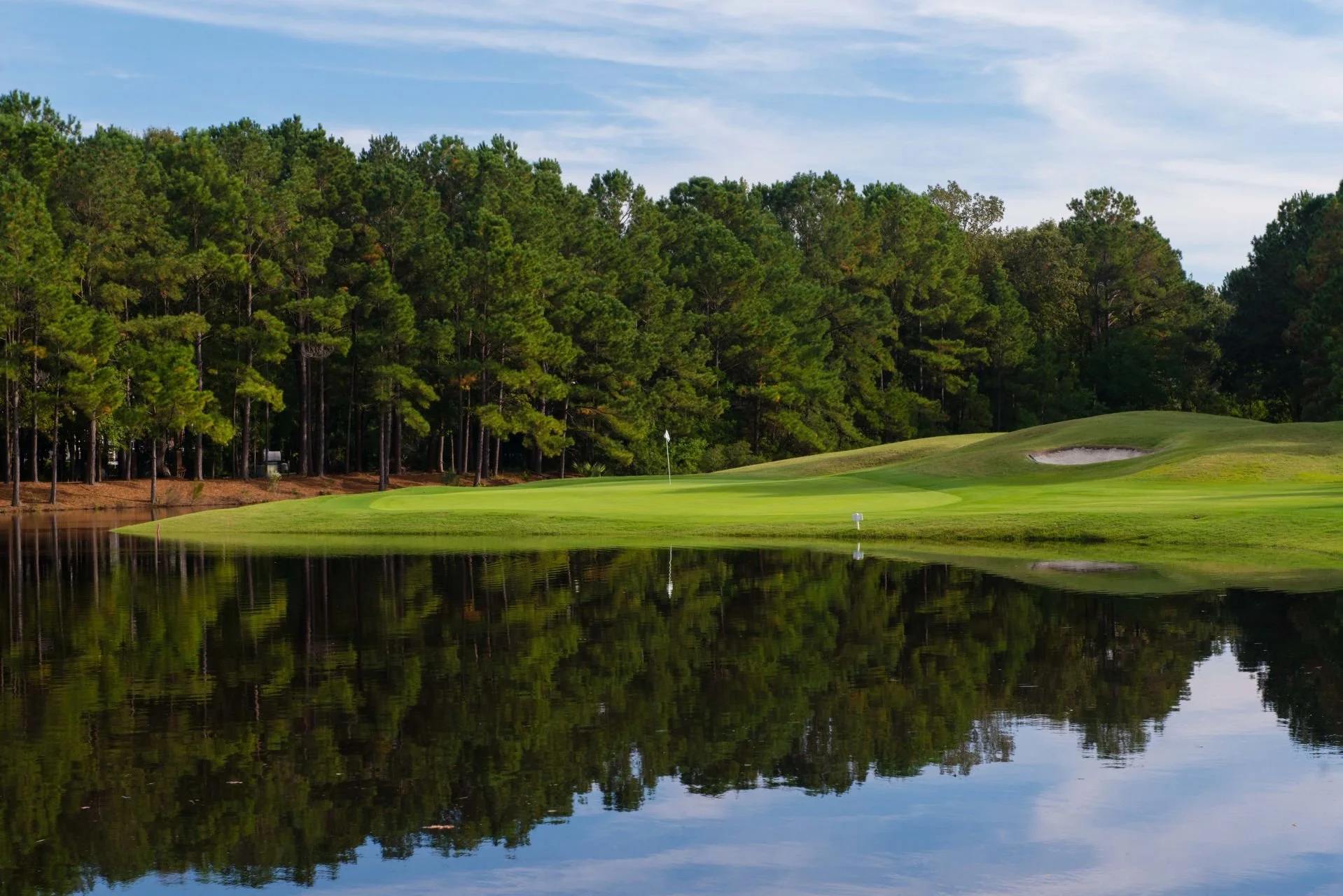 A golf course with a putting green, a flagstick, sand trap, surrounding trees, and a water hazard reflecting the landscape under a partly cloudy sky.