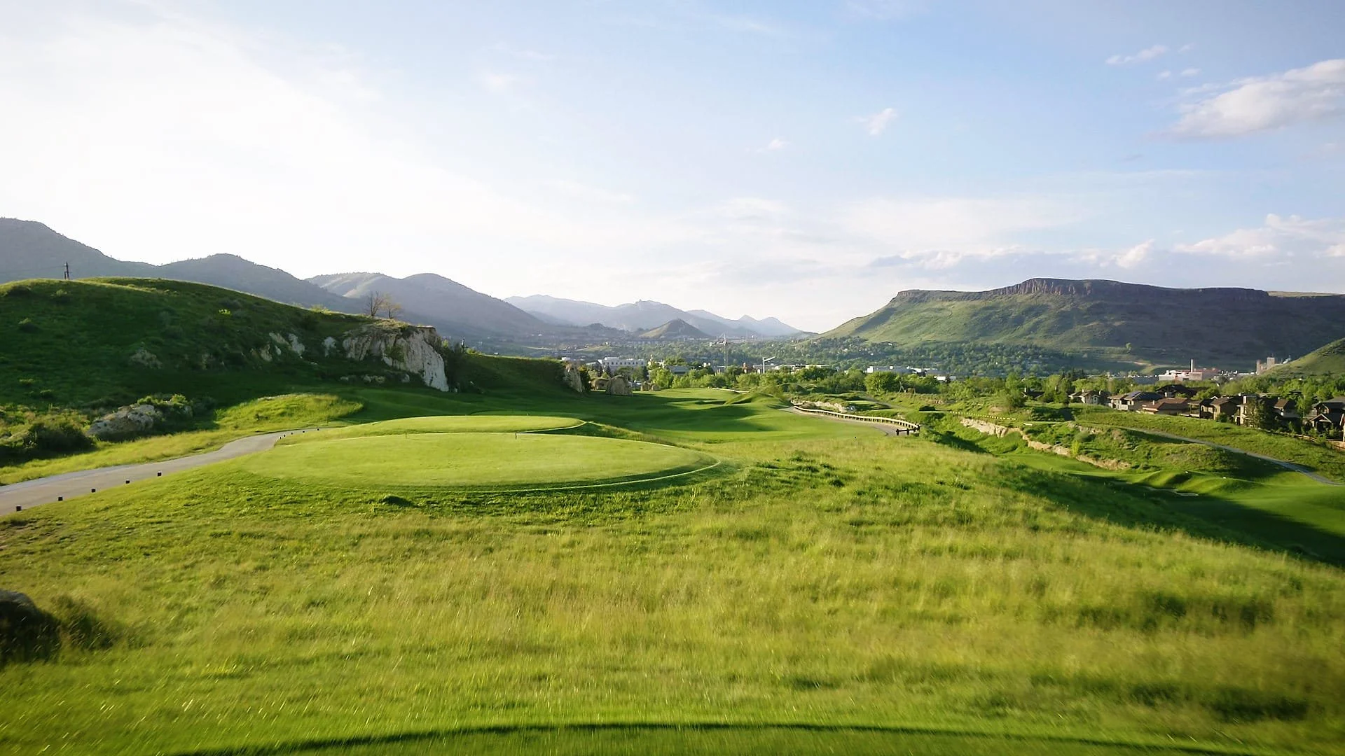 Golf course with green fairways and hills, mountains in the background, under a partly cloudy sky.