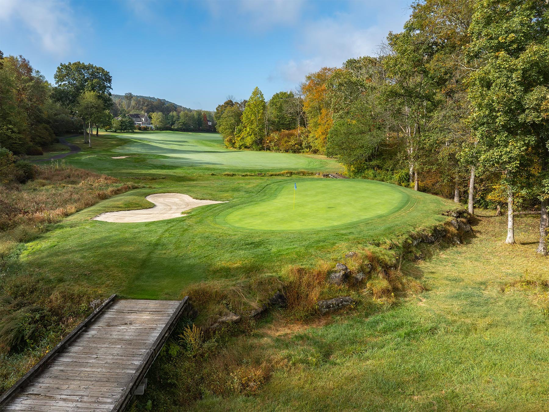 A golf course with a green and a hole marked by a flag, surrounded by trees and a small wooden bridge in the foreground.