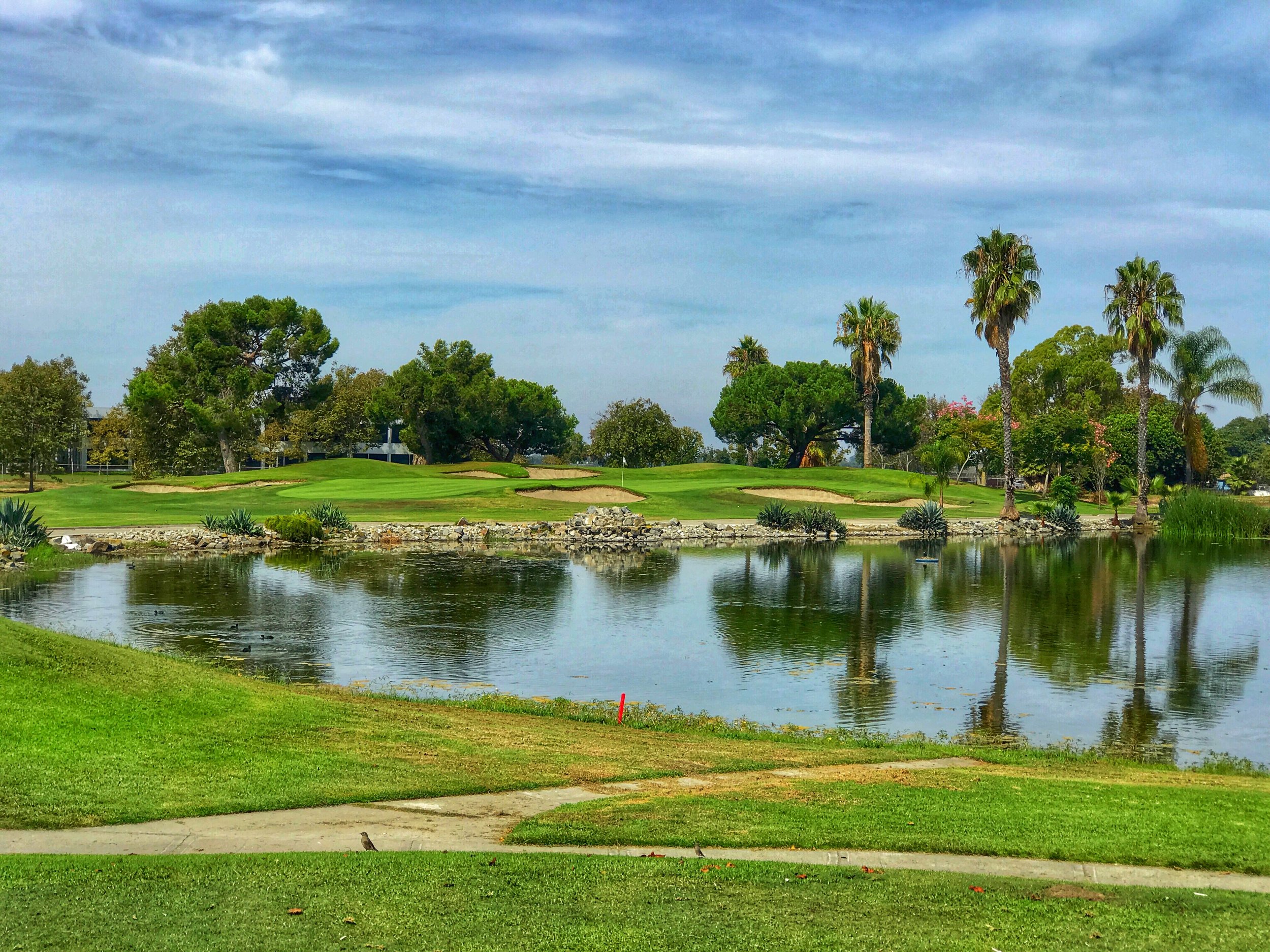 A golf course with manicured green grass, sand traps, colorful trees, and palm trees beside a reflective water hazard under a partly cloudy sky.