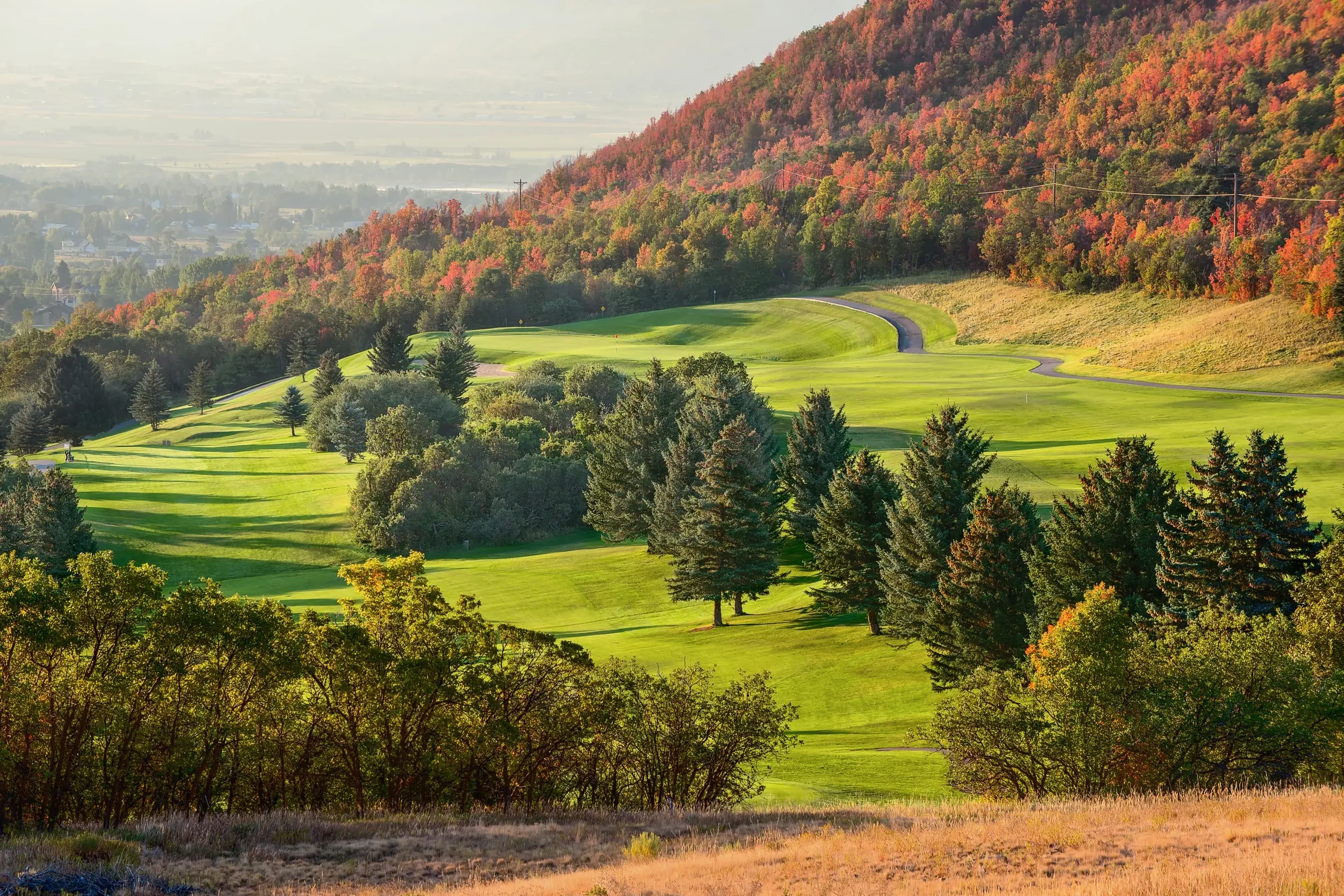 A vibrant golf course with bright green fairways and tee boxes, surrounded by lush trees and a hillside with colorful fall foliage, under a hazy sky.