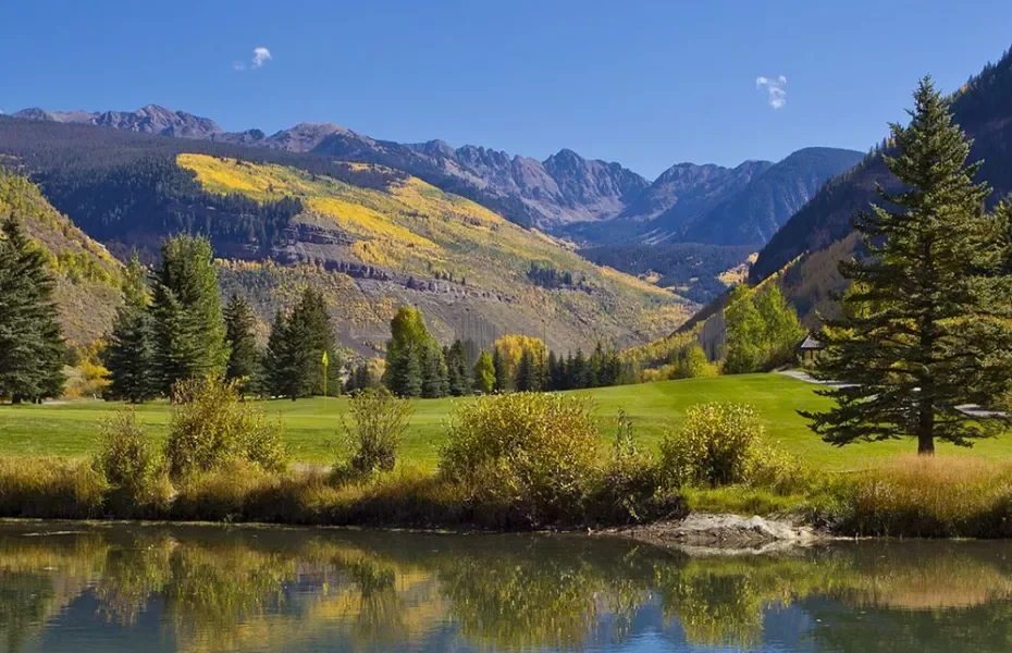 Scenic landscape of rolling green hills with a small pond in the foreground, tall pine trees, and mountains with patches of yellow foliage in the background on a clear day.