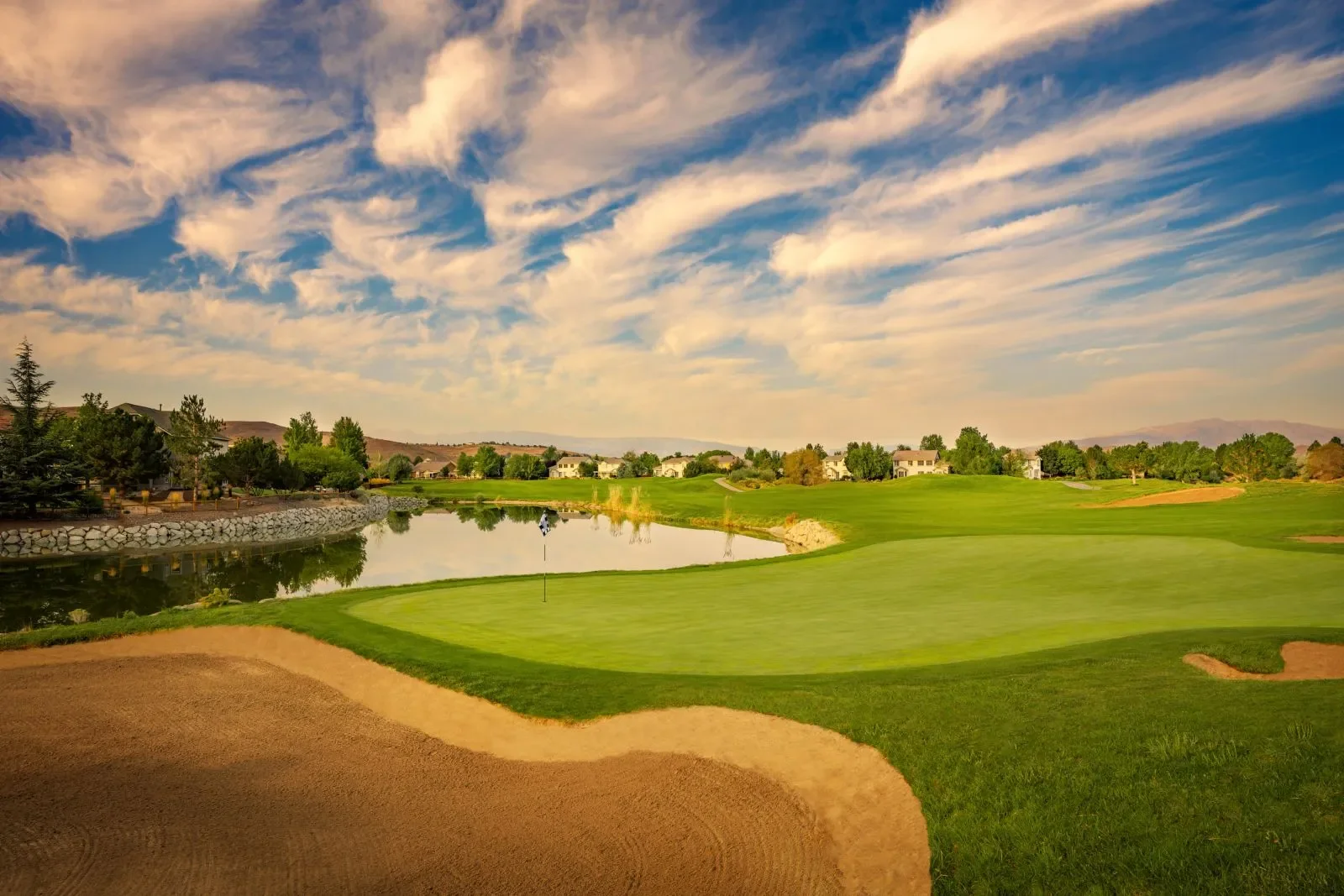 A golf course with a green, sand traps, a water hazard, and a few houses in the background under a partly cloudy sky.