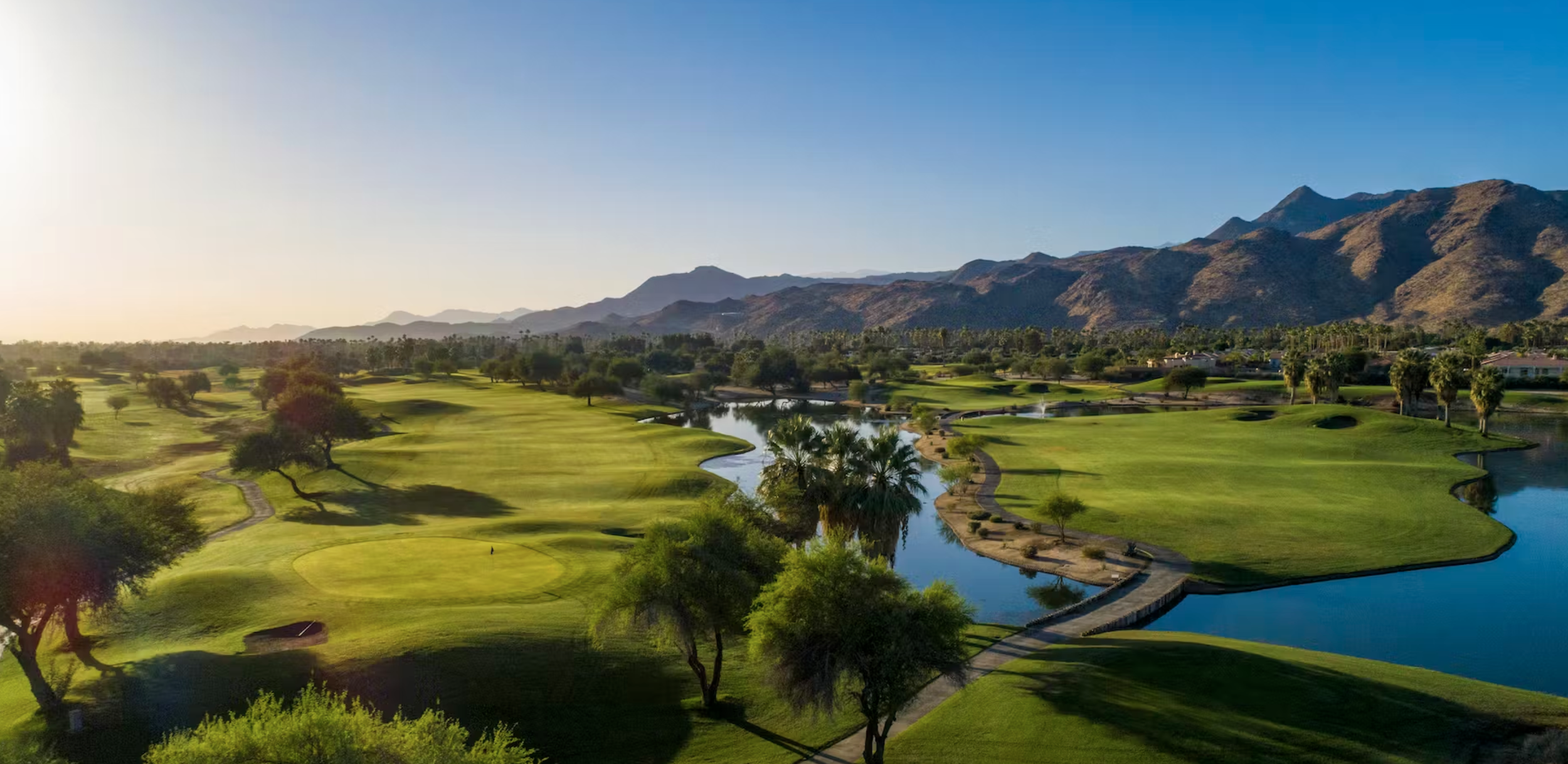 A scenic golf course with green fairways, a water hazard, palm trees, and mountains in the background under a clear blue sky.