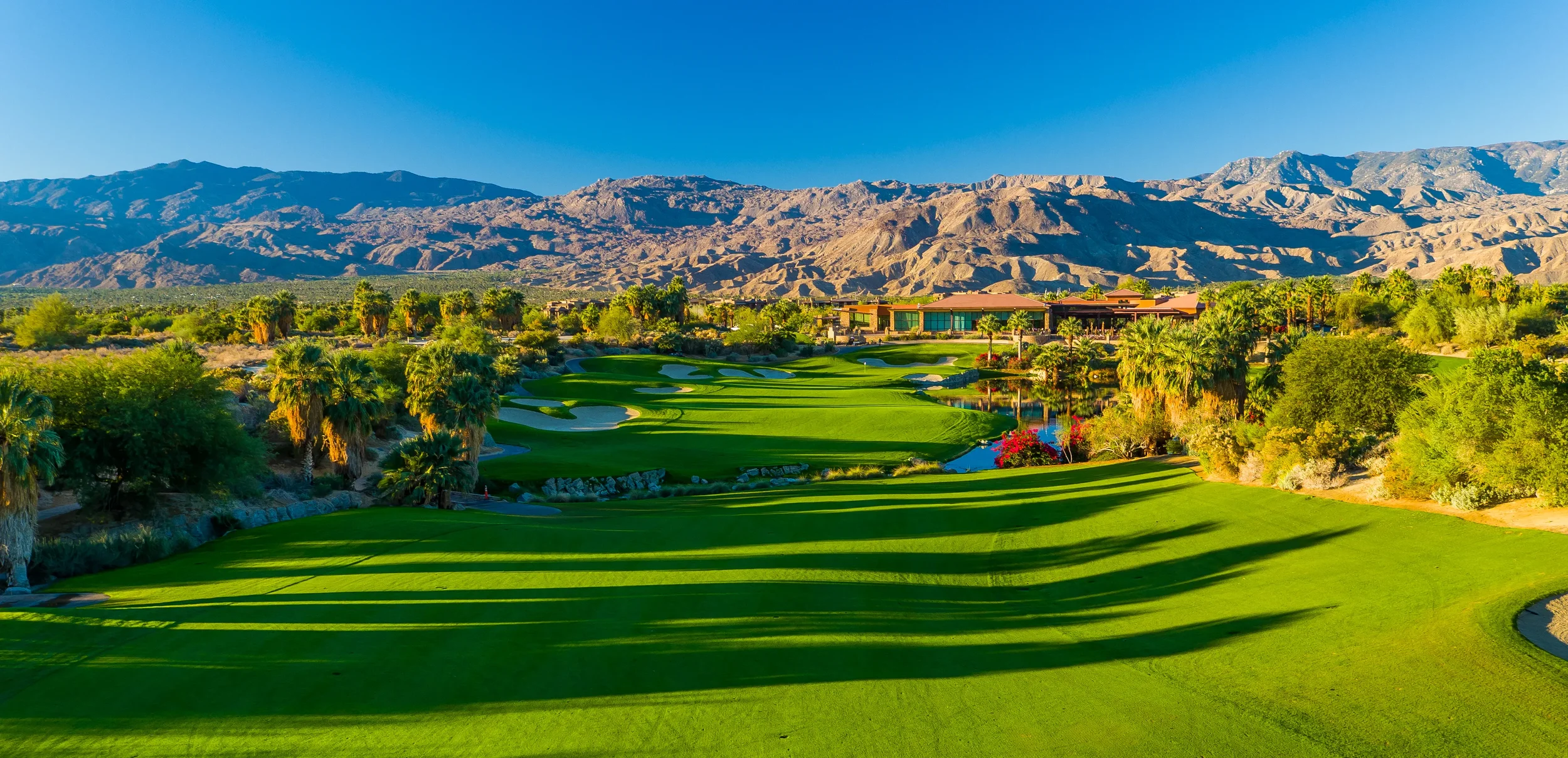 Golf course with lush green fairways, palm trees, and sand bunkers, set against a mountainous desert landscape under a clear blue sky.