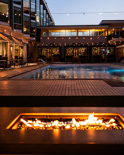 An outdoor swimming pool area at night with string lights overhead and a fire feature in the foreground
