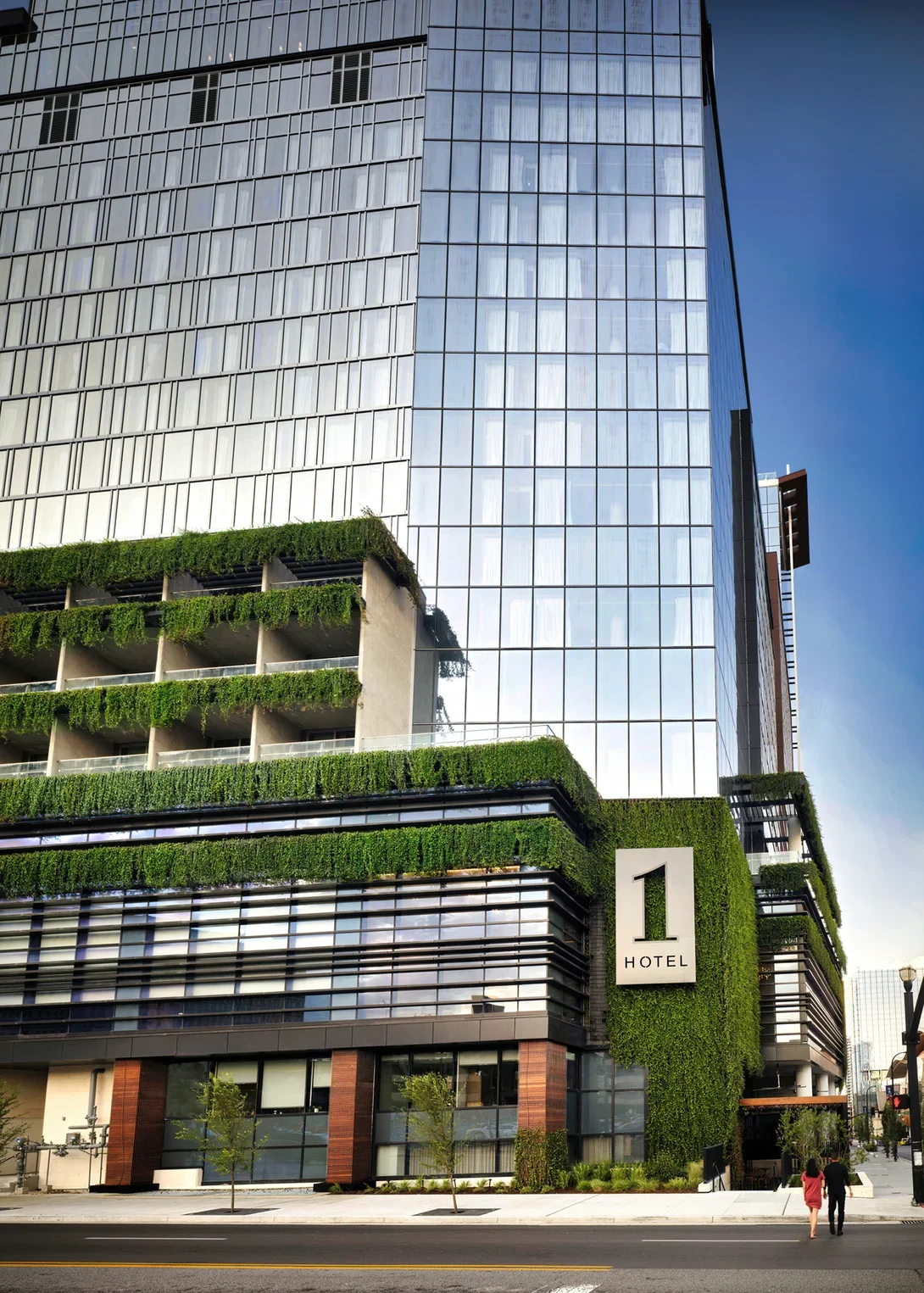 Modern hotel building with glass facade and greenery on balconies near the street with pedestrians.