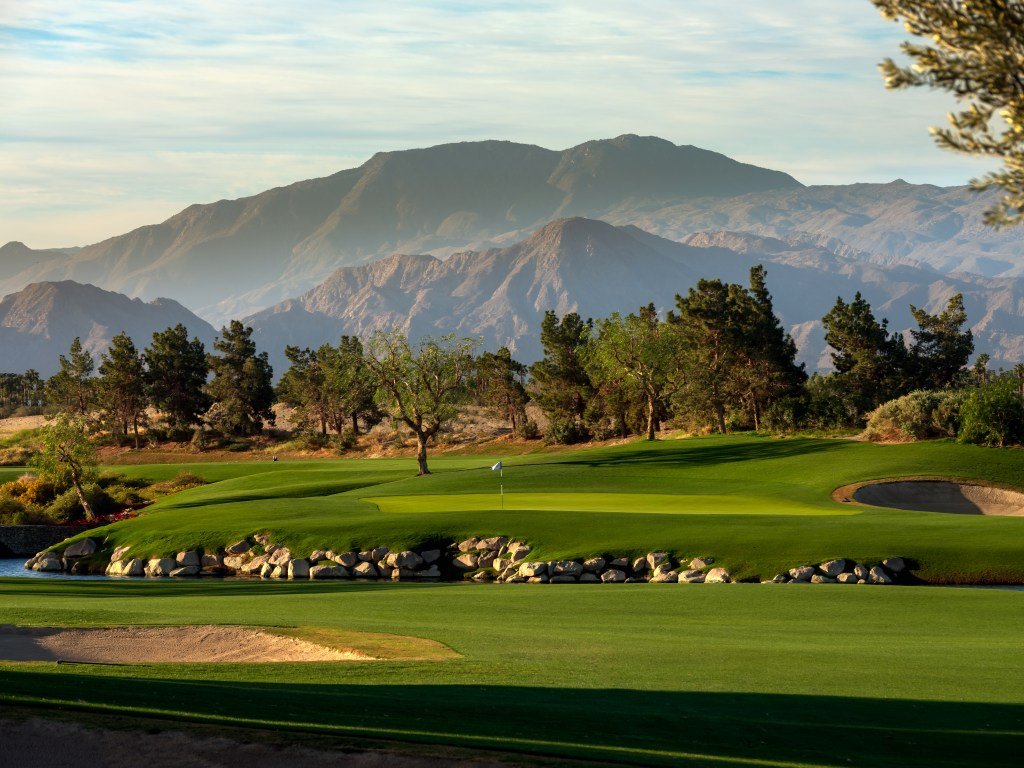 A scenic golf course with a green, sand trap, and trees, set against a backdrop of mountains under a partly cloudy sky.