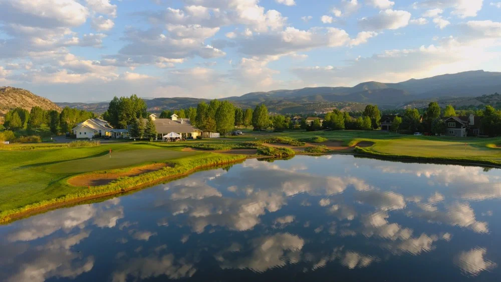A scenic view of a golf course with green fairways and a water hazard reflecting the sky and clouds, with residential houses and mountains in the background.