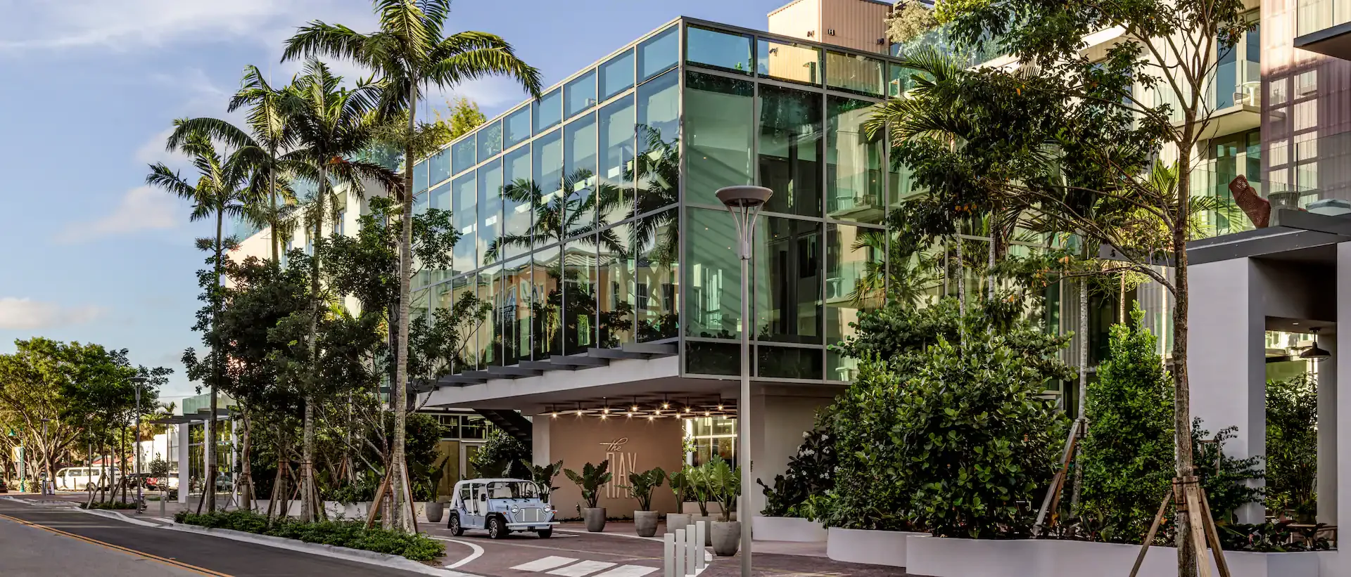 Modern glass building surrounded by lush trees and plants, with a sidewalk and street in front, a small white vintage vehicle parked underneath, and a streetlamp in the foreground.
