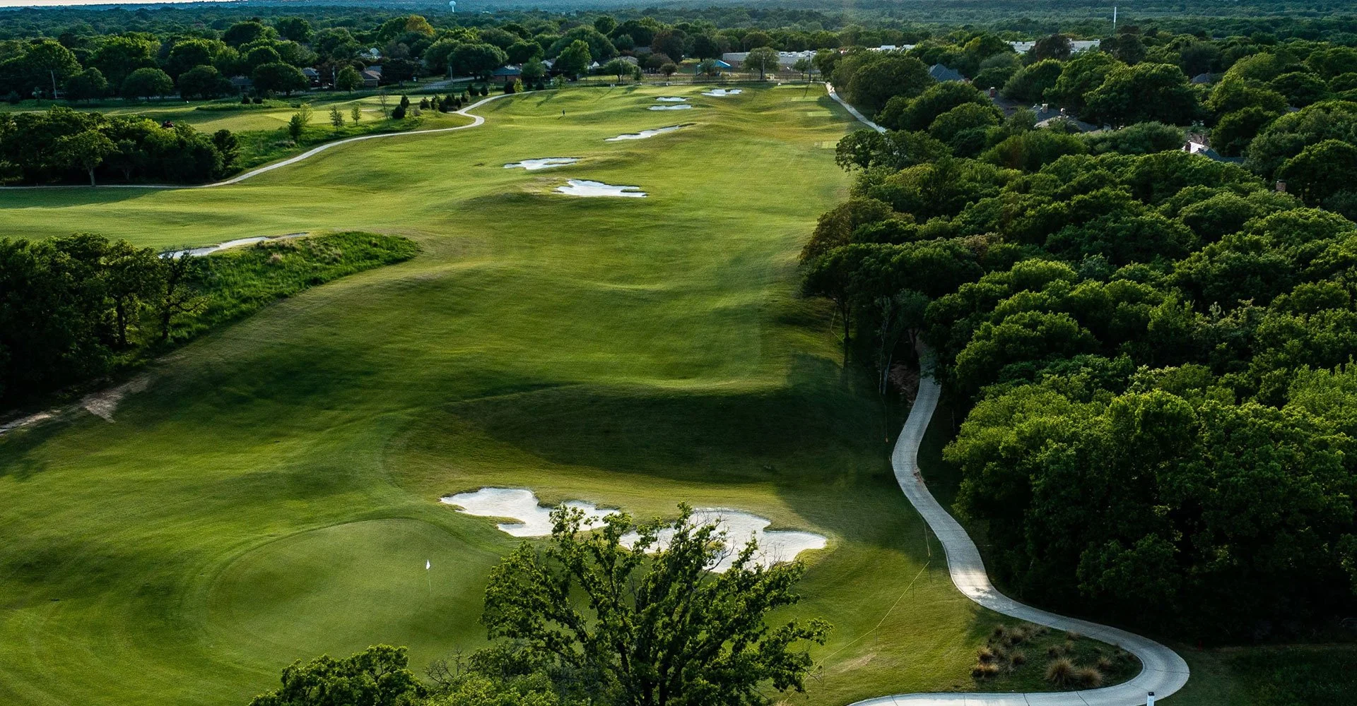 Aerial view of a golf course with lush green fairways, sand bunkers, and a paved walking path winding through the course, surrounded by dense trees, with houses and a water tower visible in the distance.