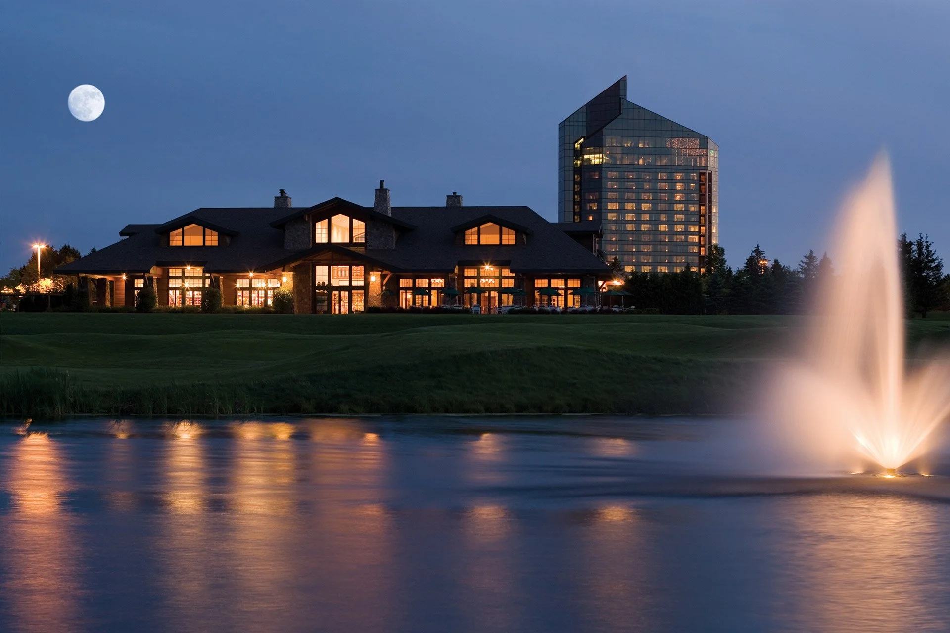 A night view of a building with lit windows, a fountain in a water body in the foreground, and a full moon in the sky.