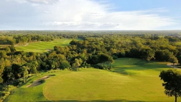 A scenic view of a golf course with lush green fairways, trees, and a partly cloudy sky in the background.