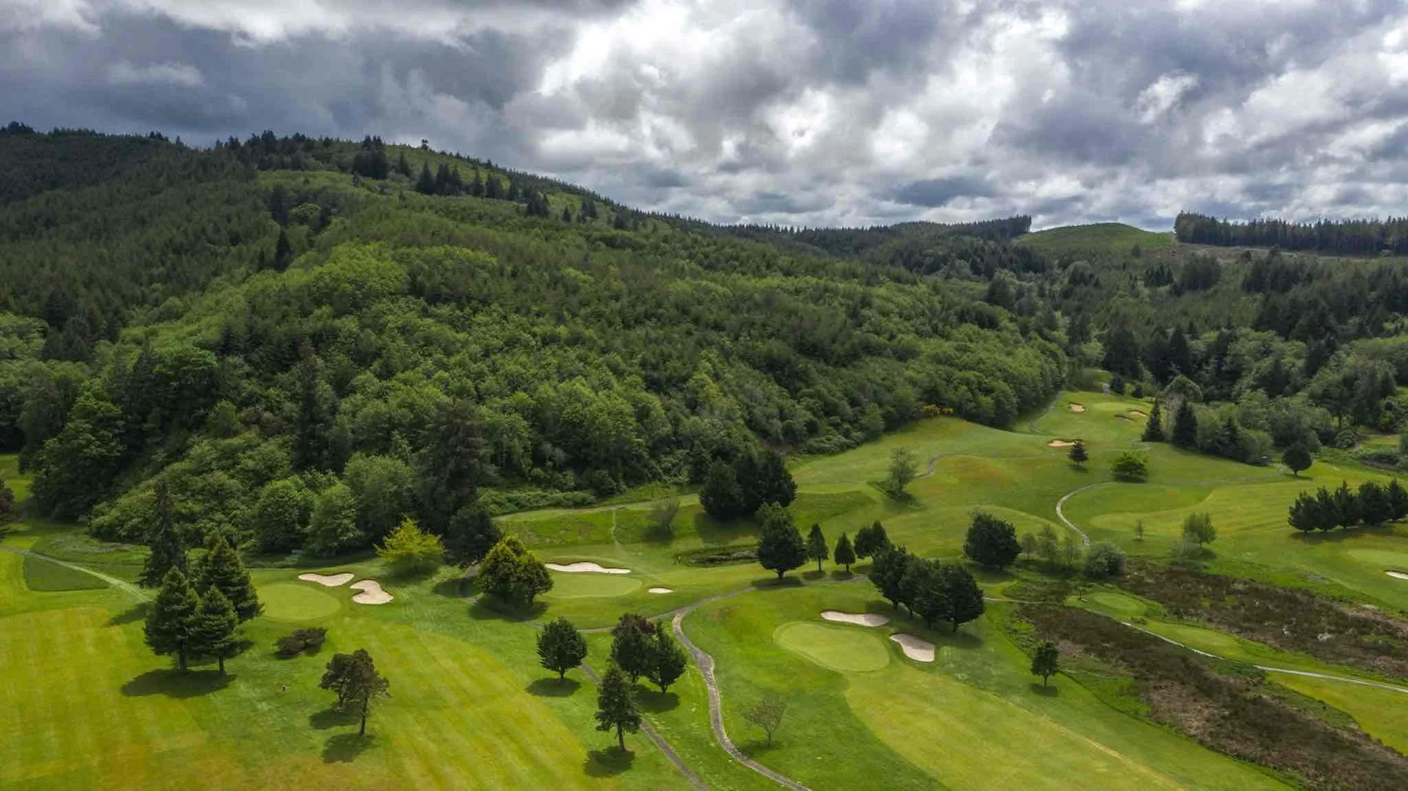 A scenic view of a golf course surrounded by lush green trees and hills, with sand bunkers and walking paths visible on a cloudy day.