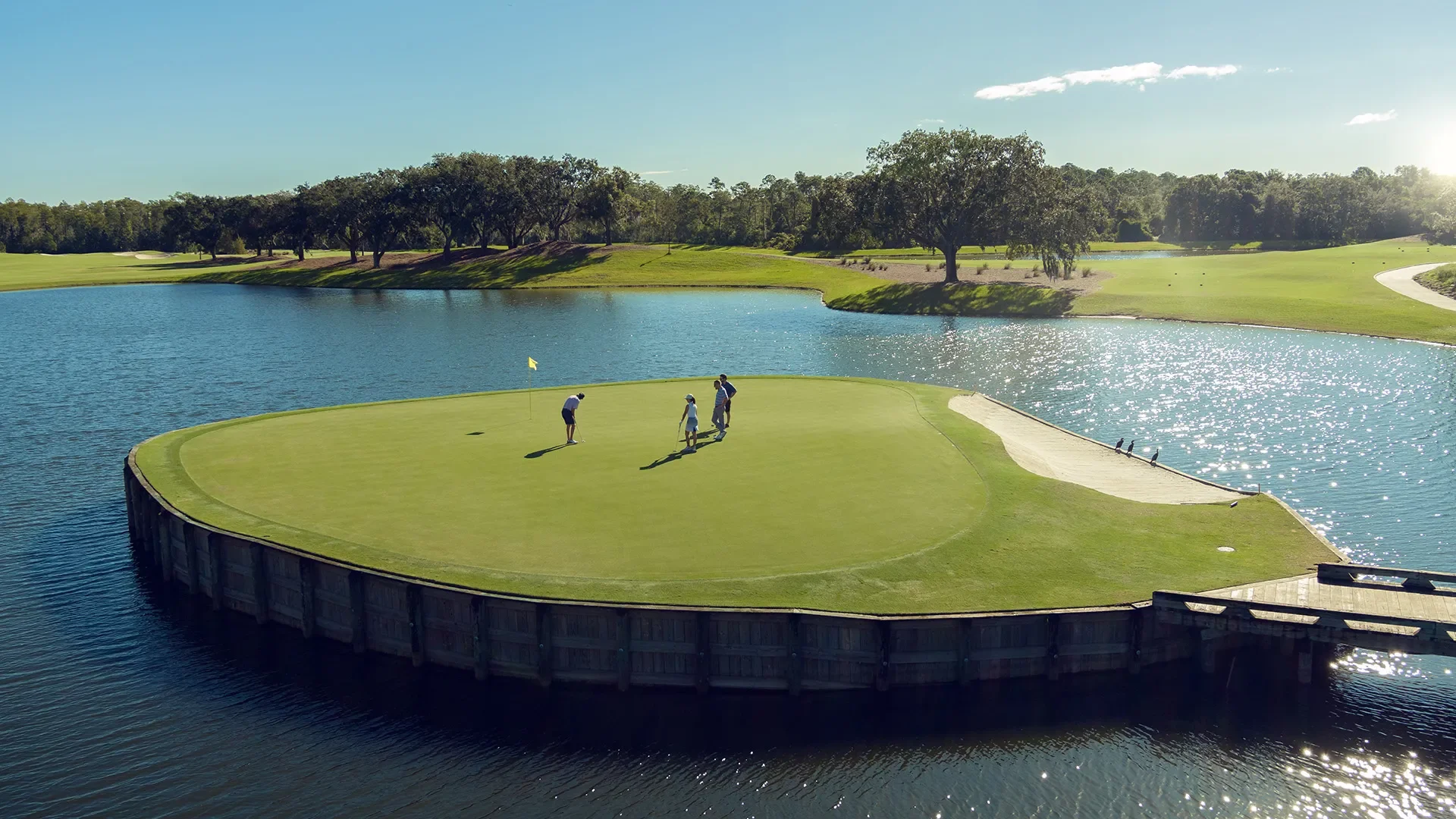 A golf course with a small green island surrounded by water, with four people playing golf on the island, and trees in the background.