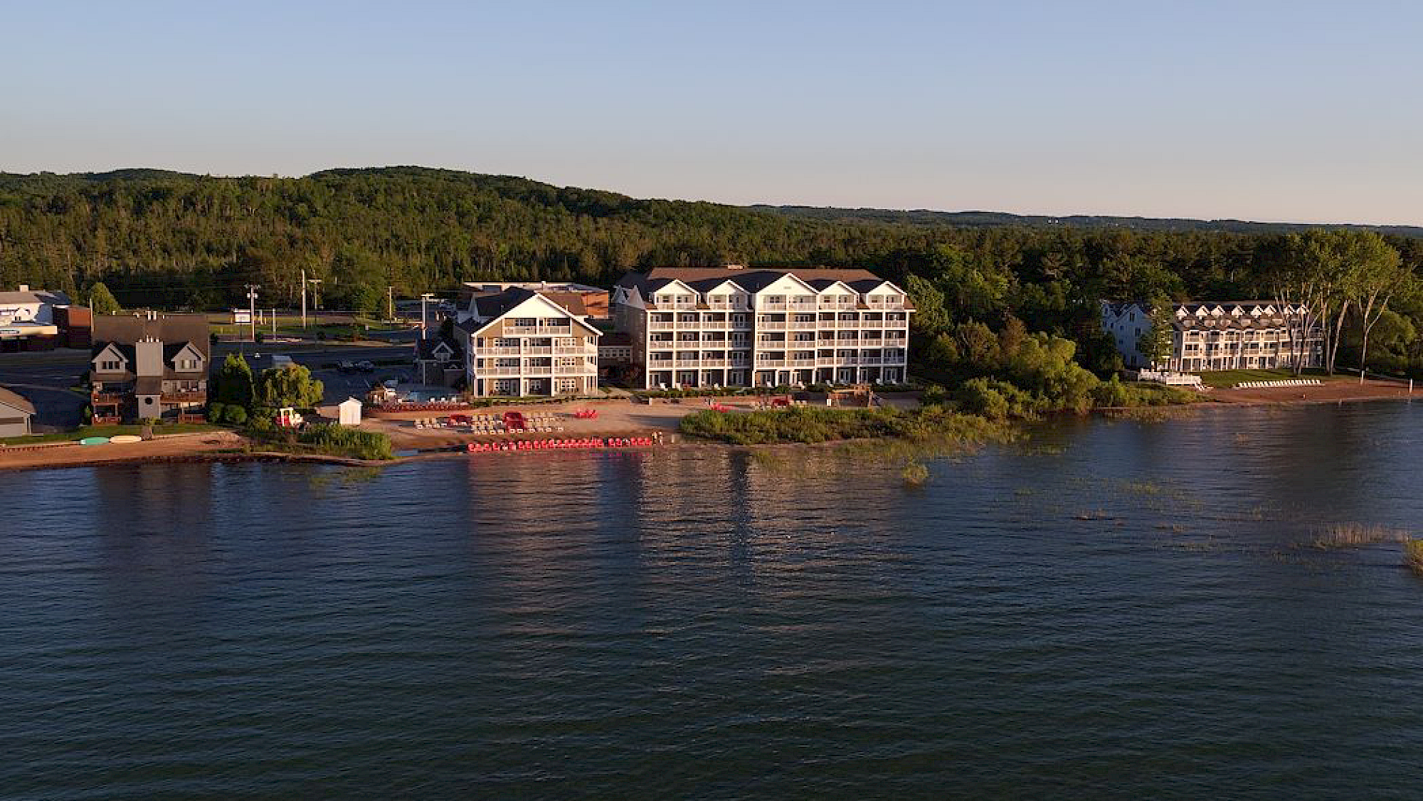 Aerial view of a lakeside resort with multiple modern white apartment buildings, small houses, a sandy beach with red amenities, and a lush green forest in the background.