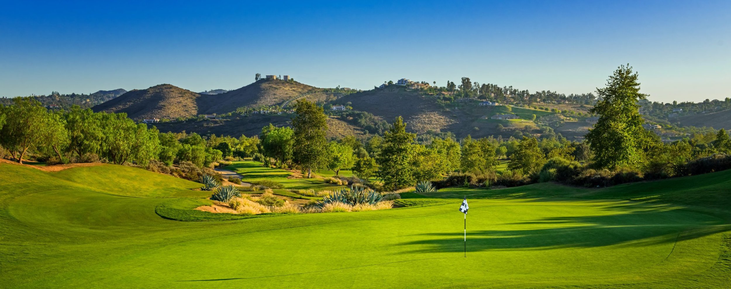 A scenic golf course with lush green grass, trees, and a flag on the putting green, with rolling hills and mountains in the background under a clear blue sky.