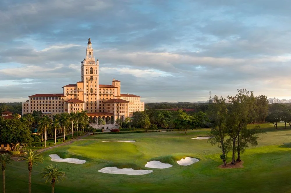 A large building resemblance of a castle or hotel next to a golf course with sand traps and trees, under a partly cloudy sky.