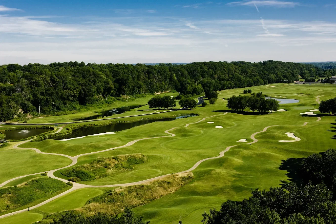 A scenic golf course with lush green fairways, sand traps, water hazards, and trees under a partly cloudy sky.