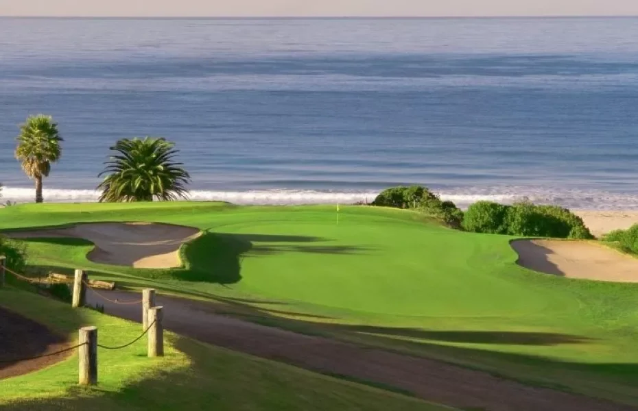 A coastal golf course with green fairways, sand bunkers, and palm trees near the ocean.