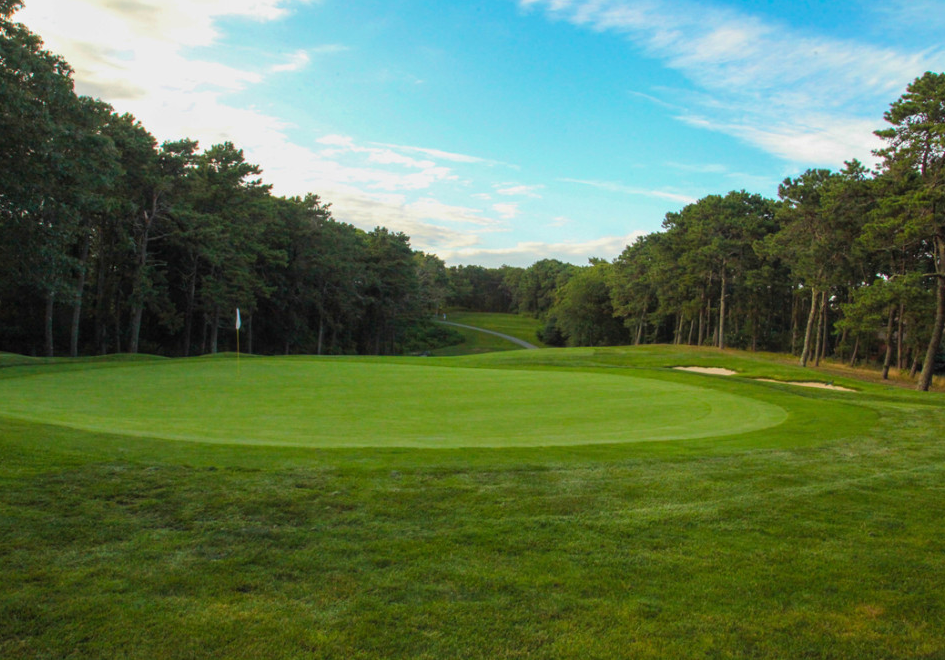 A scenic golf course with lush green grass, a flagstick on the putting green, sand bunkers, and trees surrounding the area under a partly cloudy sky.