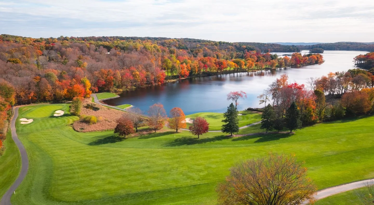 An aerial view of a golf course with green fairways and sand traps near a large lake surrounded by fall-colored trees.
