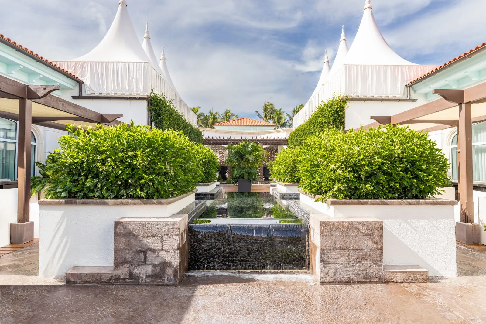 Luxury resort with white tent-like structures, green bushes, water feature, and palm trees in background.