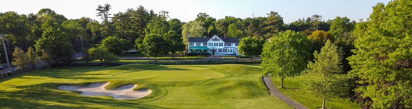 A golf course with a putting green, sand bunkers, a paved pathway, and a large white house with a porch, surrounded by green trees.