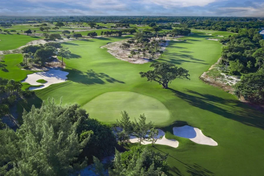 Aerial view of a golf course with lush green fairways, sand bunkers, and scattered trees under a partly cloudy sky.