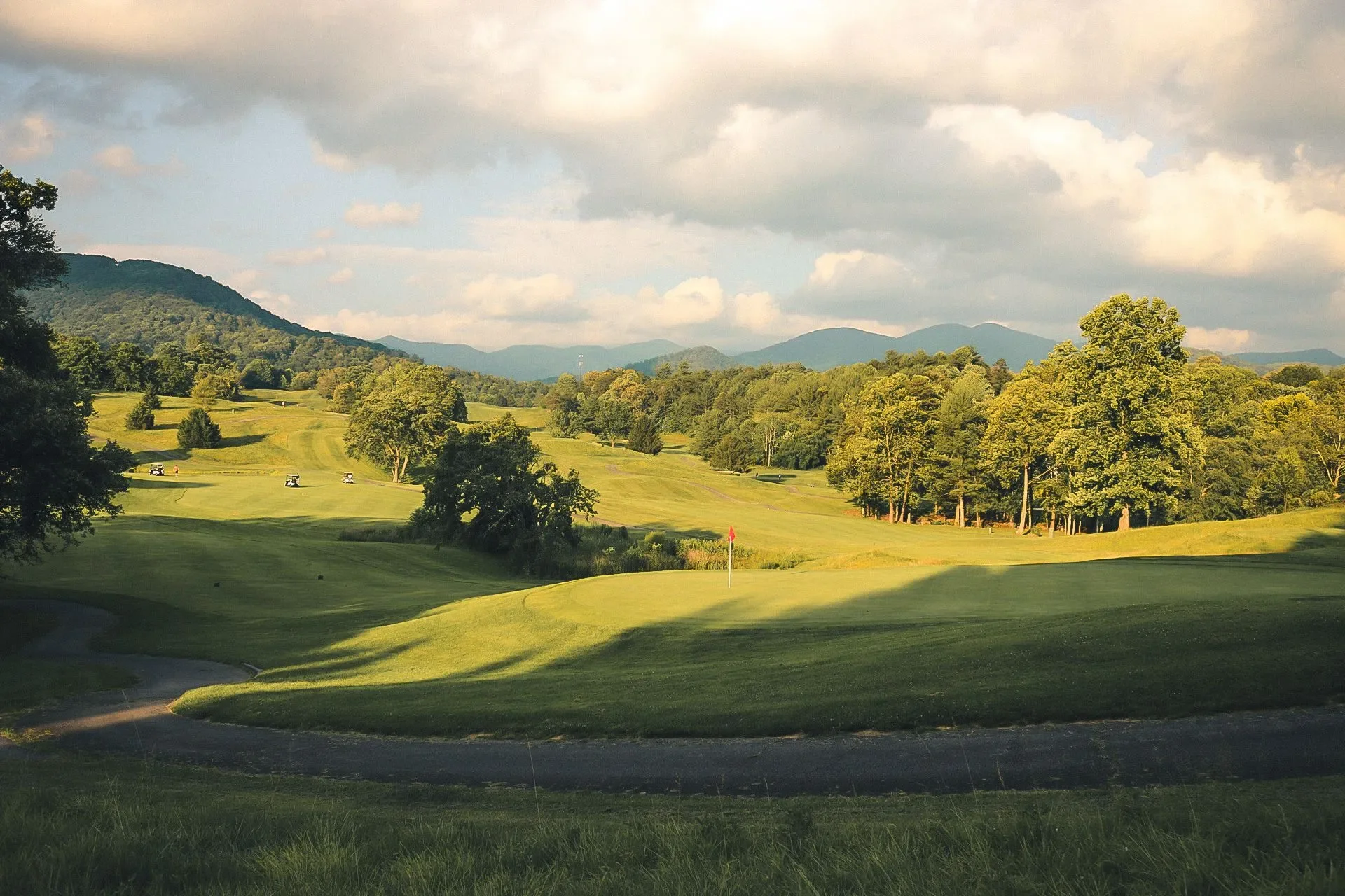 A scenic golf course with lush green fairways, scattered trees, golf carts in the distance, and a mountainous landscape in the background under partly cloudy skies.