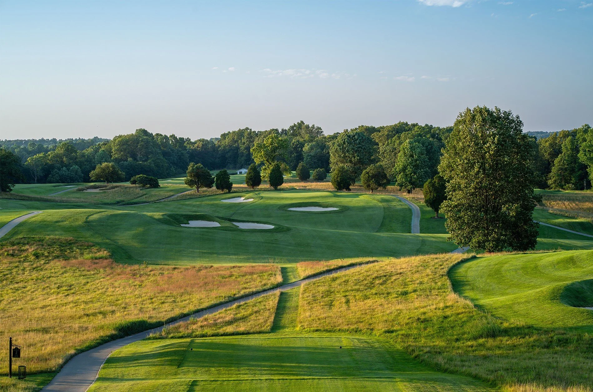 A scenic view of a golf course with lush green fairways, sand traps, and scattered trees under a clear blue sky.