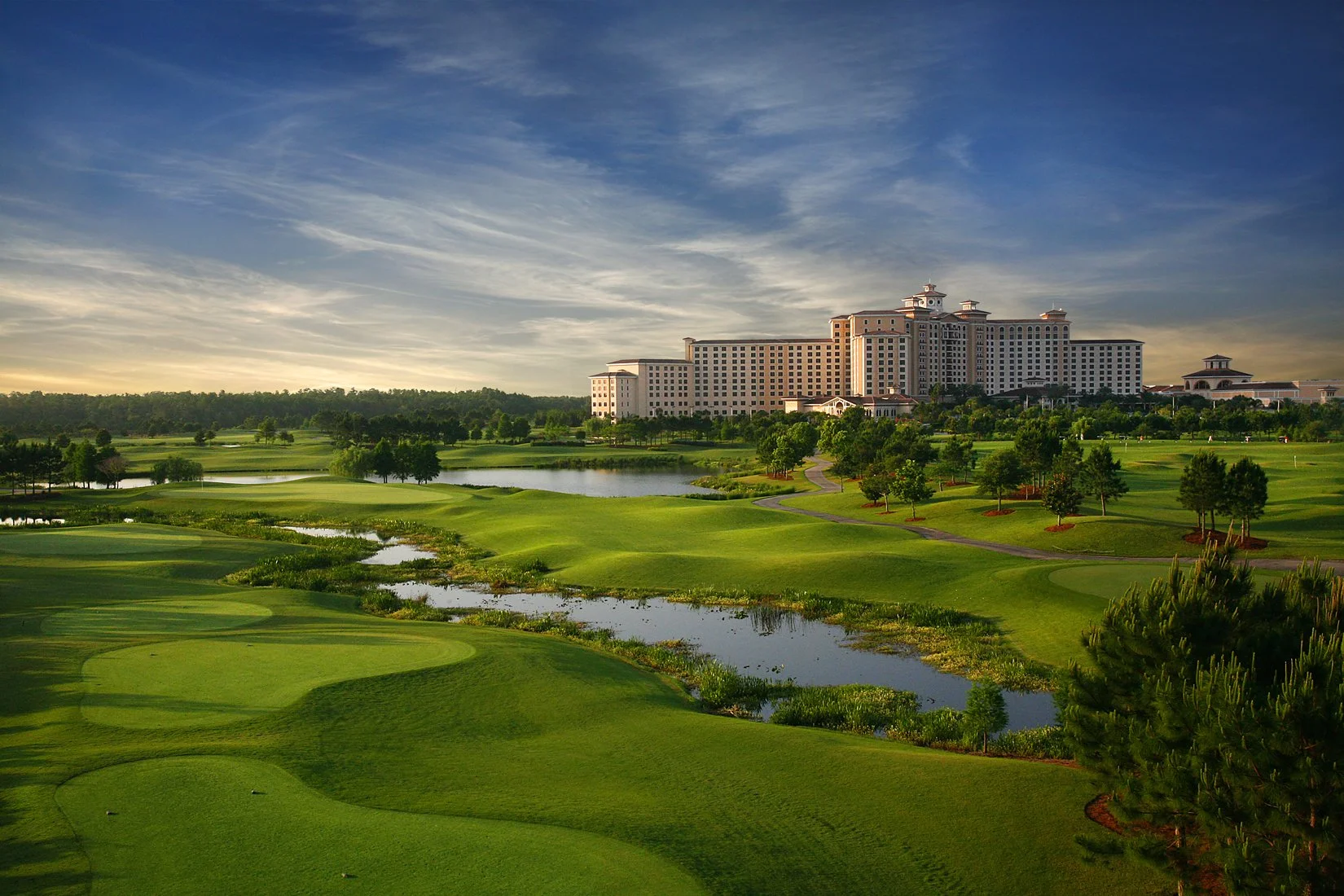 A golf course with neatly mowed greens, water hazards, and trees, with a large hotel or resort building in the background under a partly cloudy sky.