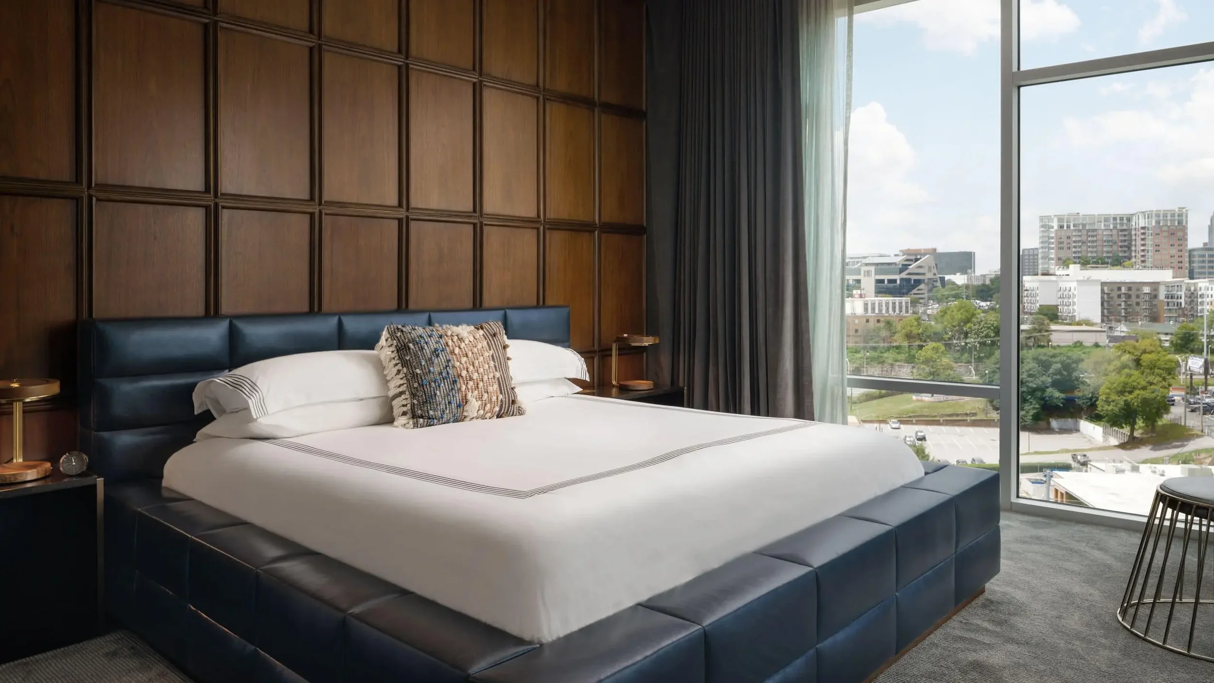 A modern hotel room with a large bed, a wood-paneled wall behind the bed, a large window with cityscape view, and a small round side table.