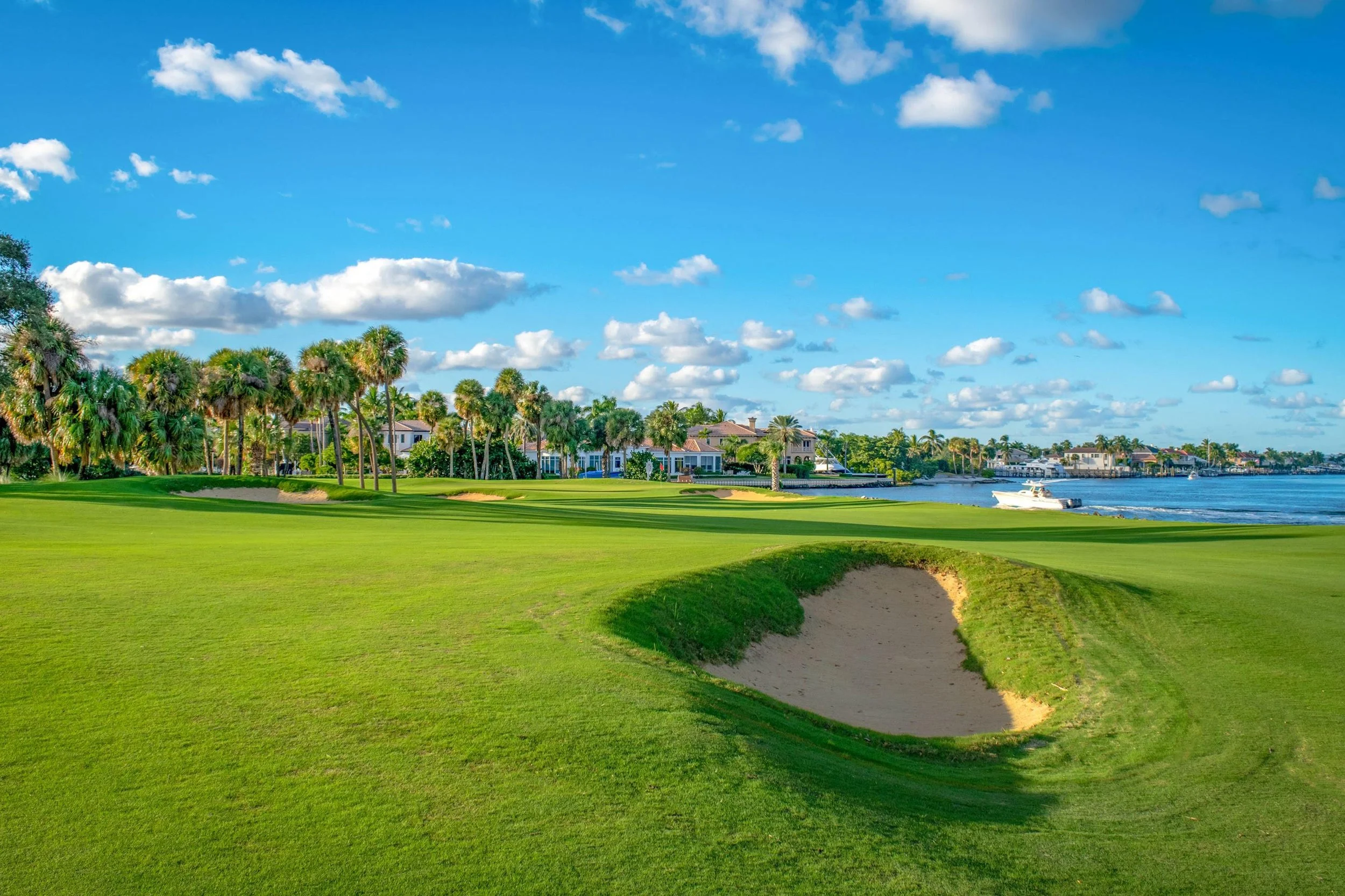 A golf course near water with sand bunkers, palm trees, houses, and a boat on the water under a partly cloudy sky.