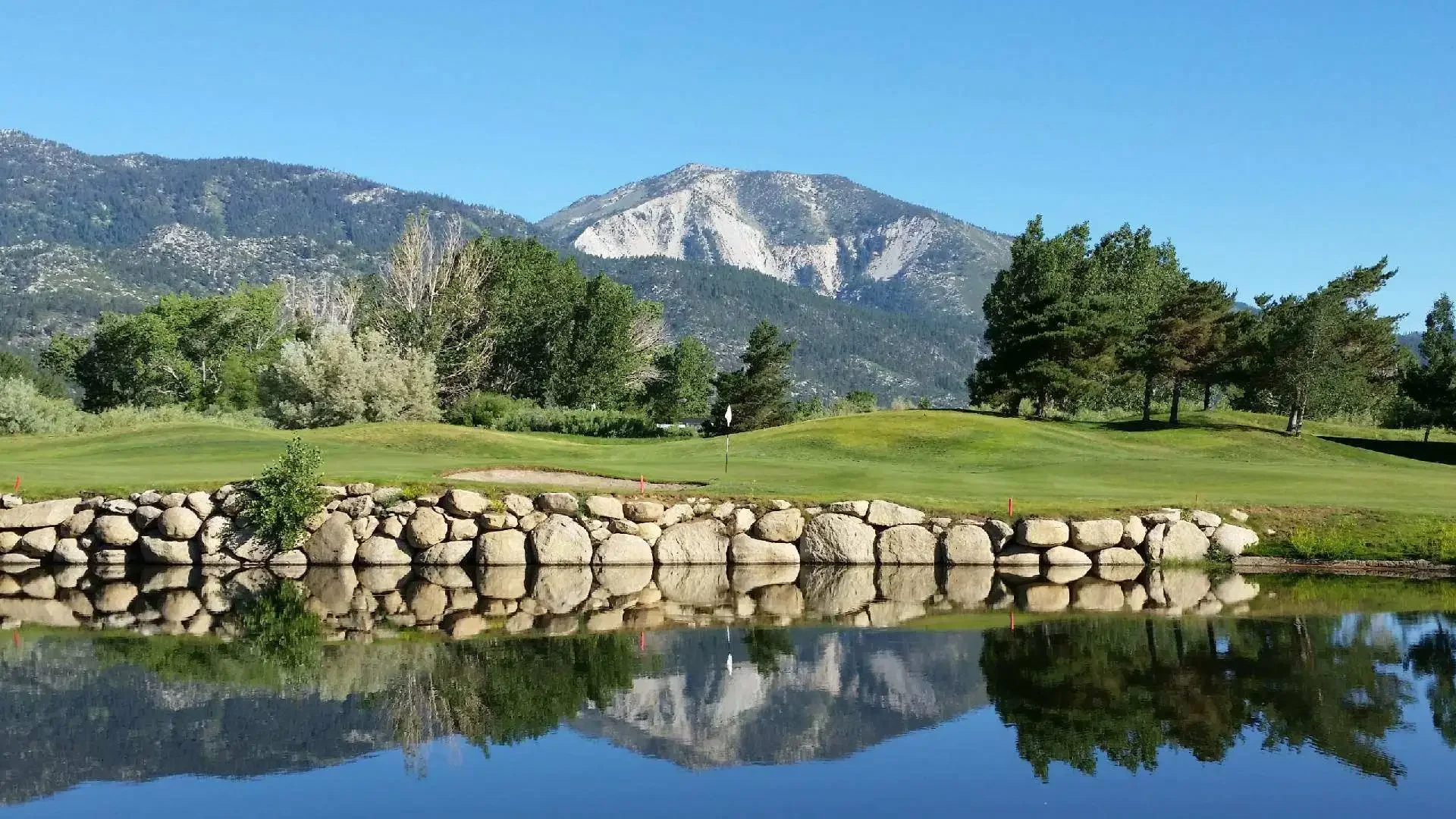Golf course with green grass, trees, stone wall, and mountain in the background under a clear blue sky, reflected in a water obstacle.