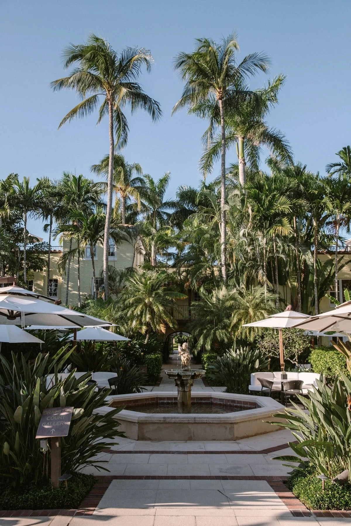 A lush tropical garden with tall palm trees, a fountain surrounded by greenery, white patio umbrellas, and outdoor seating in a resort or hotel courtyard.