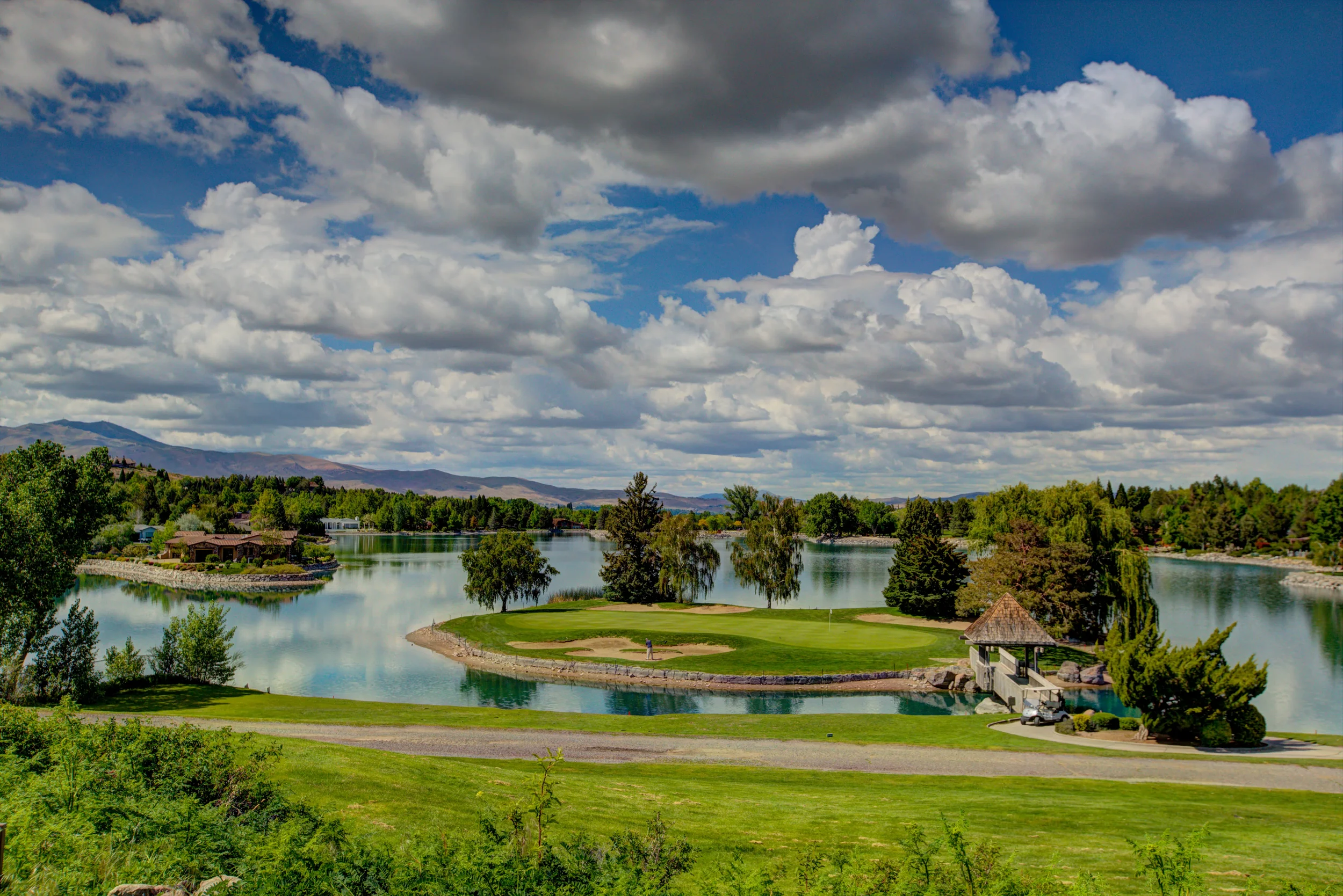 Scenic view of a golf course with a small island in the water, a golfer, a golf cart, and a gazebo, surrounded by lush green trees and hills under cloudy sky.