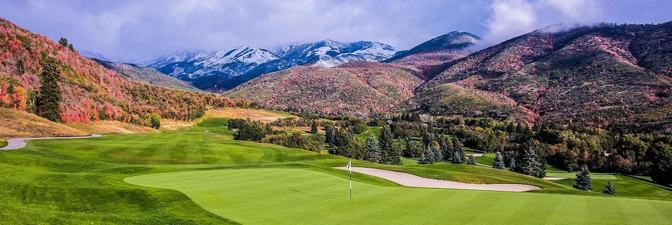 A scenic golf course with green grass, with mountains in the background covered partially with snow and colorful trees in fall.