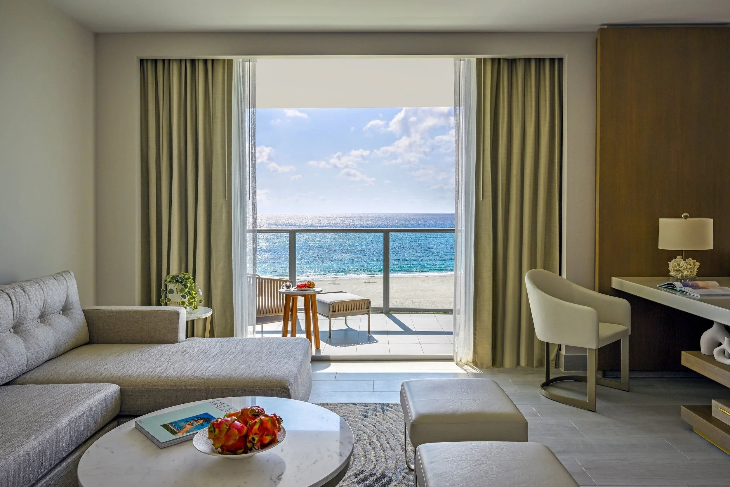 Living room with a view of the ocean, featuring a beige sofa, a round coffee table with a magazine and a bowl of apples, a small sitting area with a bench and a table on the balcony, and a desk with a lamp and chair, with curtains framing the balcony door.