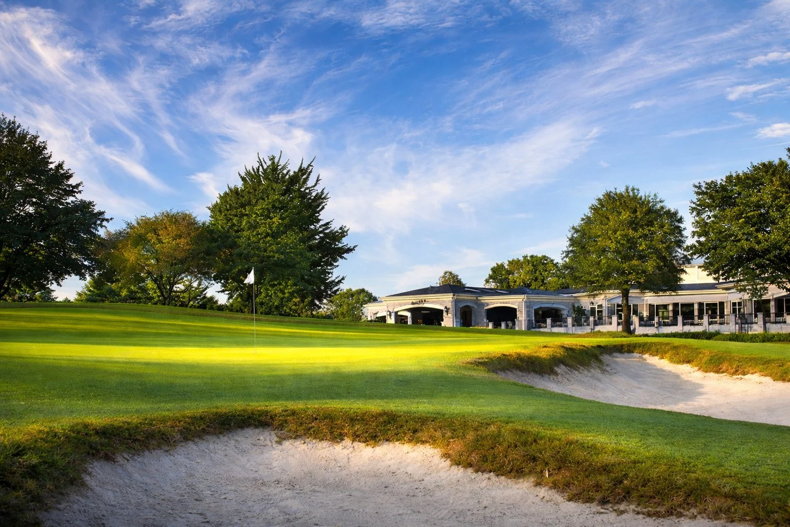 A golf course with green grass, a sand trap, and a flag on the green, with a clubhouse and trees in the background under a blue sky with scattered clouds.