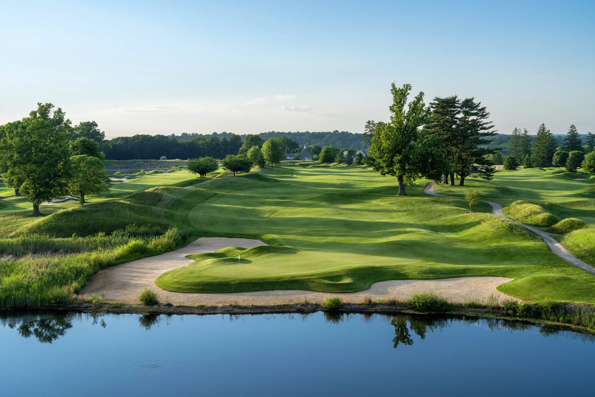 A scenic view of a golf course with lush green fairways, trees, sand traps, a water hazard in the foreground, and a clear blue sky.