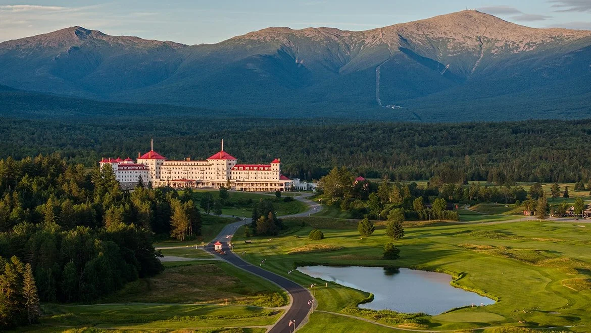 A grand hotel with red roofs and cream-colored walls surrounded by a lush golf course, with mountains in the background.