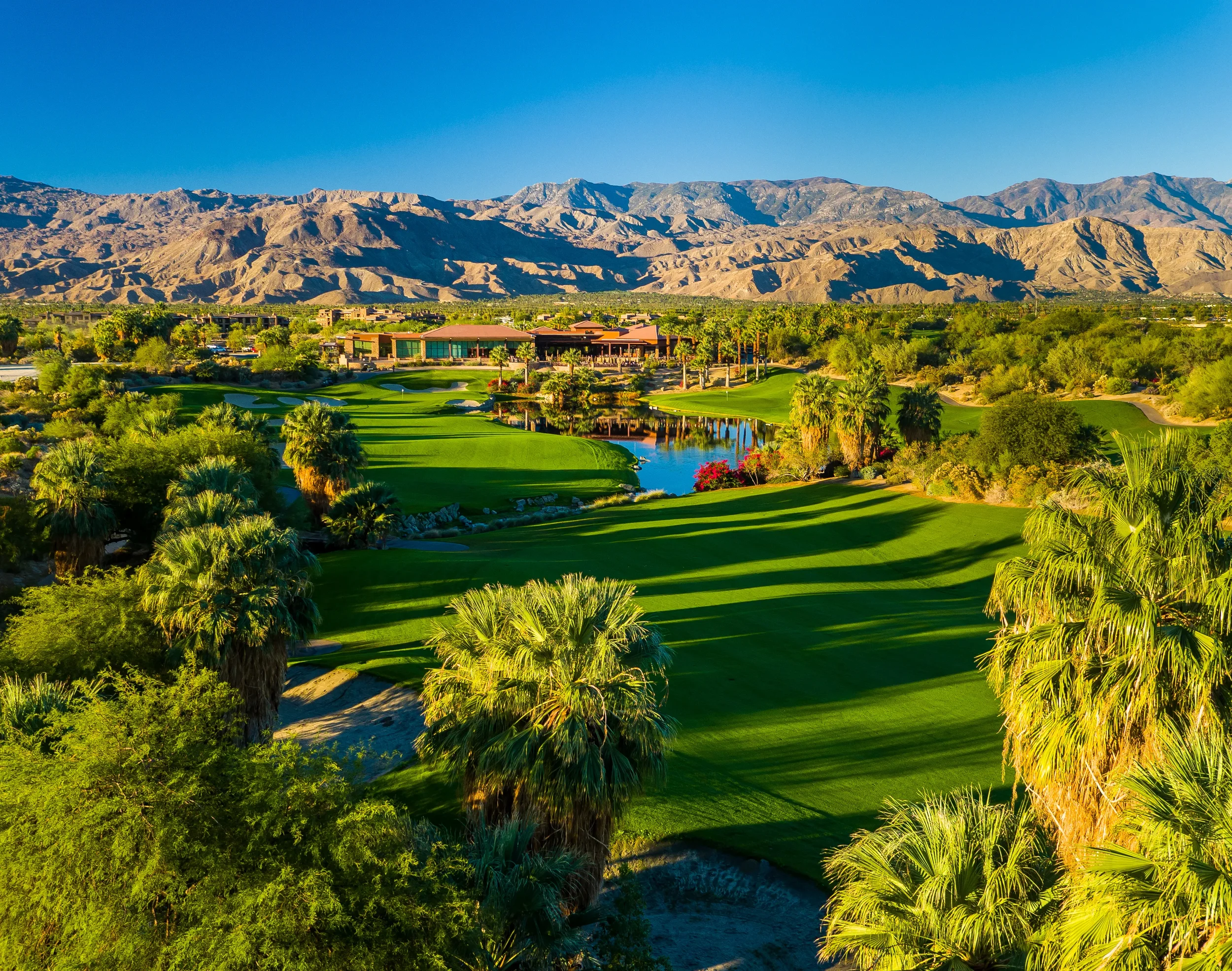 A scenic view of a lush golf course with green fairways, palm trees, water features, and a clubhouse at the background, with mountains under a clear blue sky.